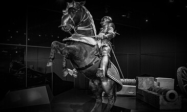 1. Black and white image of medieval knight in shining armor riding a horse with armor, displayed in a museum setting.