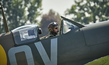Pilot in cockpit of a vintage military aircraft with helmet and sunglasses, surrounded by a blurred green tree background.