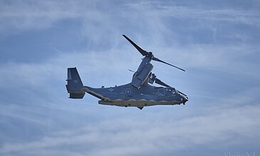 Stunning aviation photography capturing a military CV-22 Osprey in flight against a bright blue sky. Aerial view of a military CV-22 Osprey flying through the sky with clouds in the background.