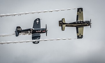 1. Two vintage Navy aircraft performing an aerial stunt with smoke trails against a cloudy sky.