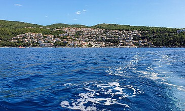 A scenic view of a hillside town along the coast, with numerous white and pastel-colored houses, lush green hills, and the sparkling blue sea under a bright sky. Vibrant coastal town with colorful houses on hillside overlooking blue waters, sunny sky, and lush green hills, capturing a picturesque seaside landscape.