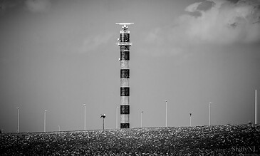 2.A black and white photo of a communication tower with a helicopter on top, surrounded by street lights on a flat ground. 1.Communication tower on a flat landscape in black and white, with cloudy sky background.