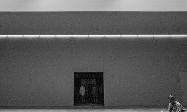 A minimalistic black-and-white image of a modern building facade with a man sitting on the sidewalk reading a book, capturing urban life and architectural beauty.
