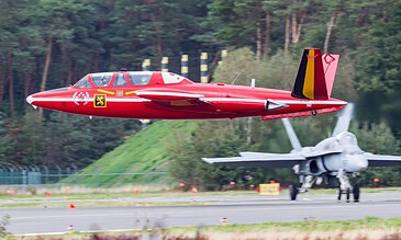 A sleek red jet aircraft taking off from the runway, surrounded by lush green trees in the background, showcasing high-speed aviation action.