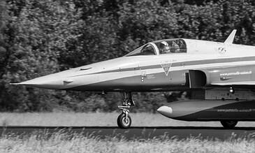 Close-up of a modern military aircraft preparing for takeoff, showcasing aerodynamic design and technical details. A sleek fighter jet on the runway with trees in the background, captured in black and white.