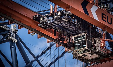 Elevated industrial crane lifting container with detailed machinery and steel structure against blue sky, highlighting moments of engineering and construction excellence. High-angle view of a large industrial crane lifting shipping containers, showcasing detailed machinery, steel structure, and blue sky background. Perfect for capturing industrial photography moments.
