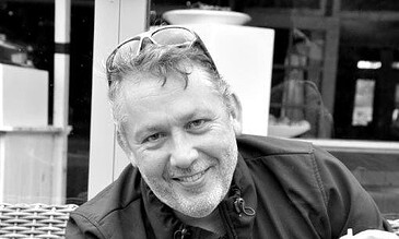 A smiling man with glasses on his head and a beard, sitting outdoors with a black dog, in a candid moment captured in black and white.