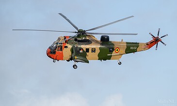 A Belgian military helicopter flying against a light blue sky with some clouds, showcasing intricate camouflage and military markings.