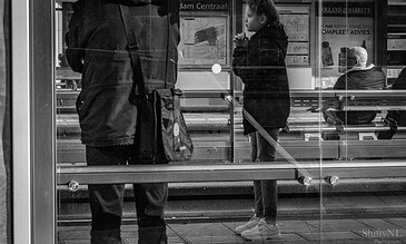 A black and white photo of a young woman waiting at a bus stop, engaged in a conversation, with other passengers seated behind her, capturing a candid moment of daily city life.