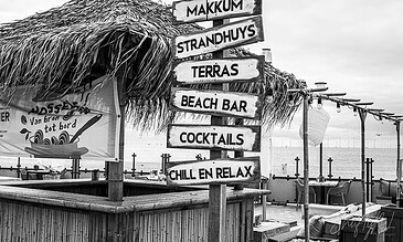 A detailed black and white image of a beach bar with multiple signs pointing to Makkum, Strandhuys, Terras, Beach Bar, Cocktails, and Chill En Relax, featuring a thatched roof and outdoor seating area.