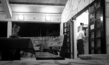 A black and white photograph of a man placing an order at a drive-thru window in an outdoor, rustic environment during nighttime. Man ordering at a drive-thru window at night in a rustic outdoor setting, black and white photo capturing a candid moment.
