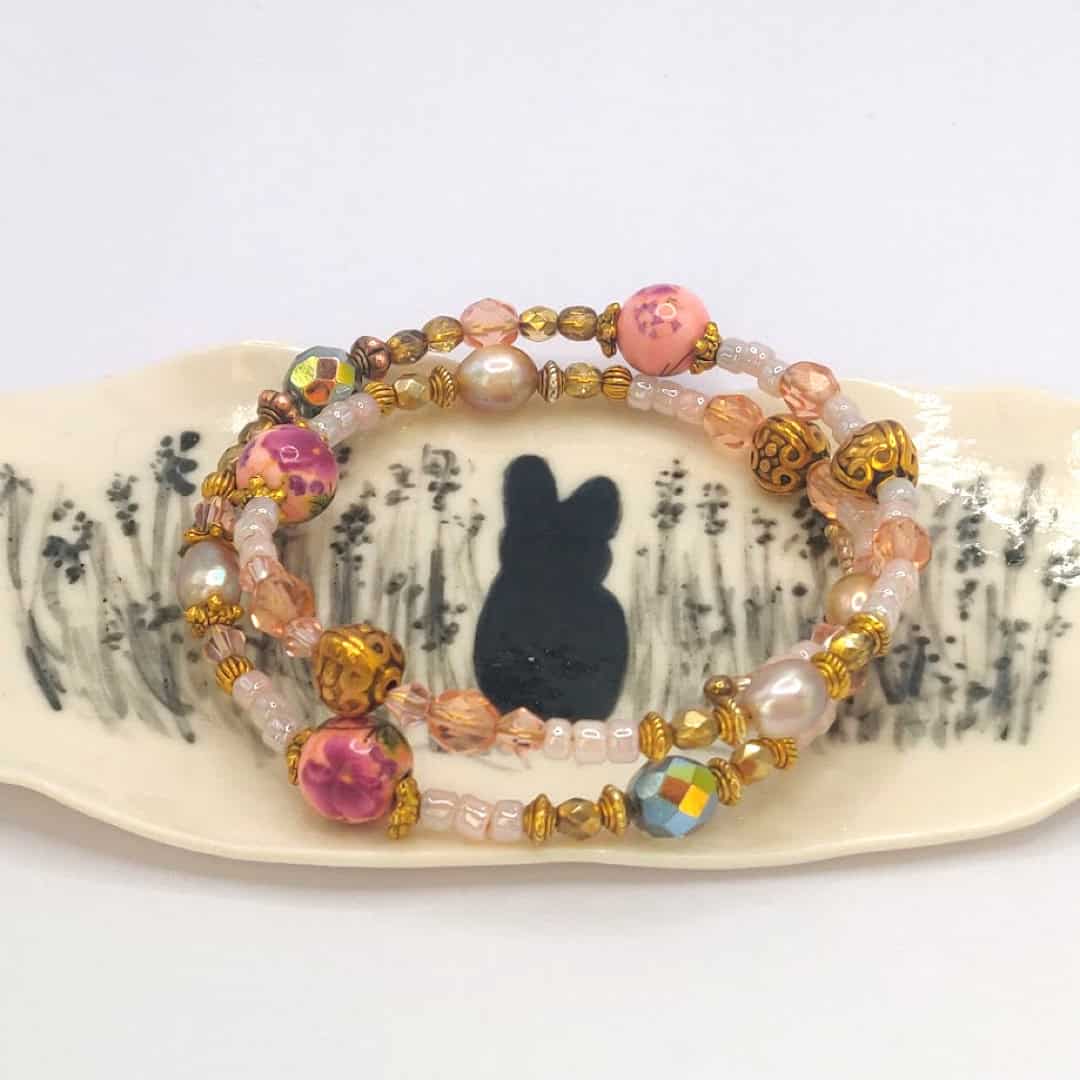 Two beaded bracelets with gold, pink, and pearl elements placed on a Bunny Jewellery Holder featuring a silhouette of a rabbit surrounded by plants.