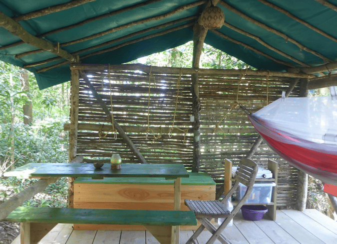 A photo of the camping accommodations at Hamac Camp in Guadeloupe. The small, rustic shelter features a picnic table and chairs as well as a hammock covered with mosquito netting.