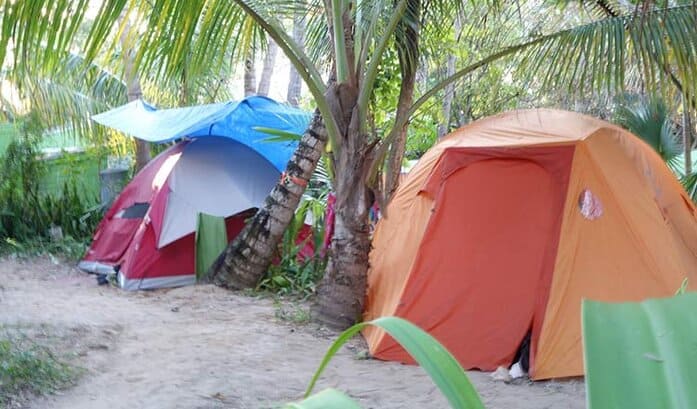 Two tents -- an orange tent, and a red and white tent with a blue tarp covering -- are pitched on a beach under lush palm trees in the Bahamas.