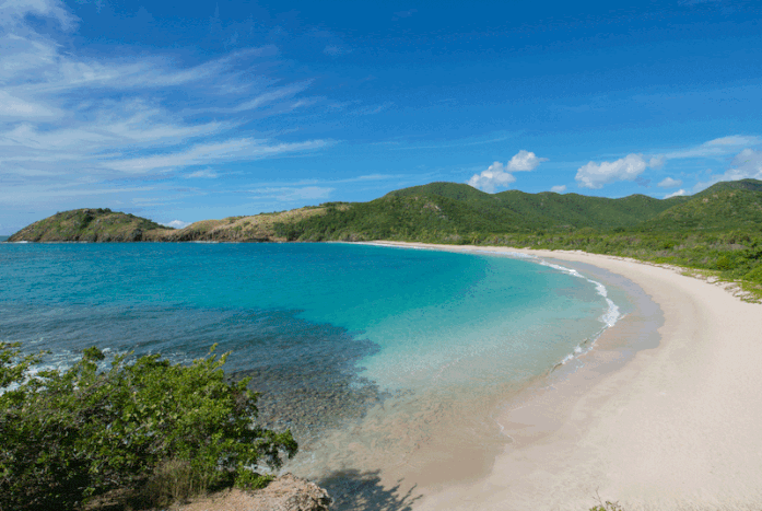 An aerial photo of Rendezvous Bay Beach in Antigua on a clear day -- the crystal blue ocean is calm, the beach is empty, and the sky is clear blue.