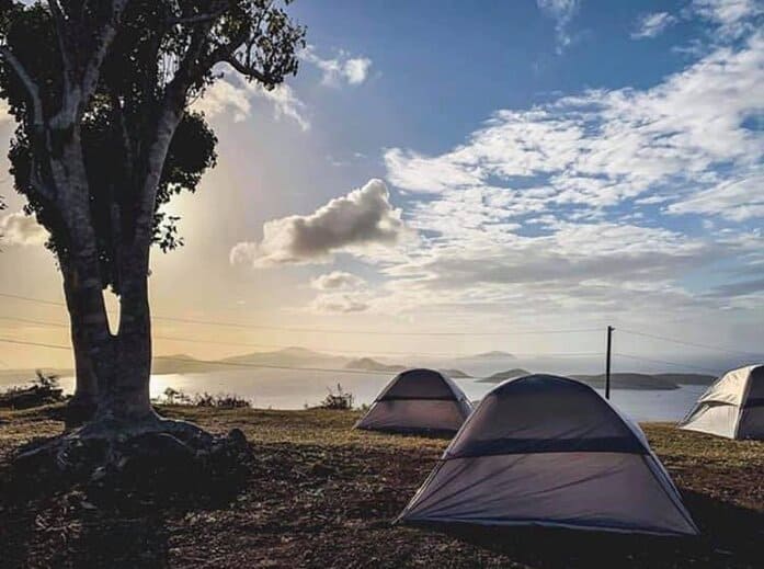 Three camping tents set up on St. John's beach in the US Virgin Islands. The sun rises over the ocean, filling a clear sky with bright light.