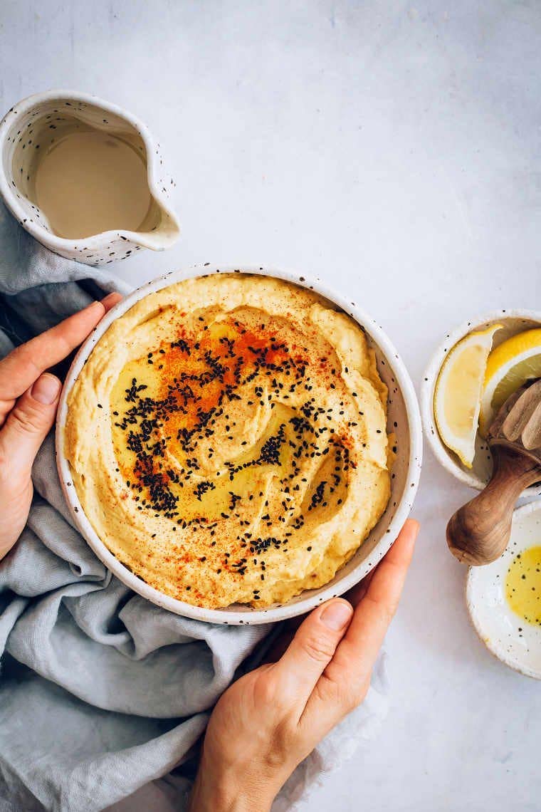 white bowl of pumpkin hummus with black seeds on a grey surface with two hands around it