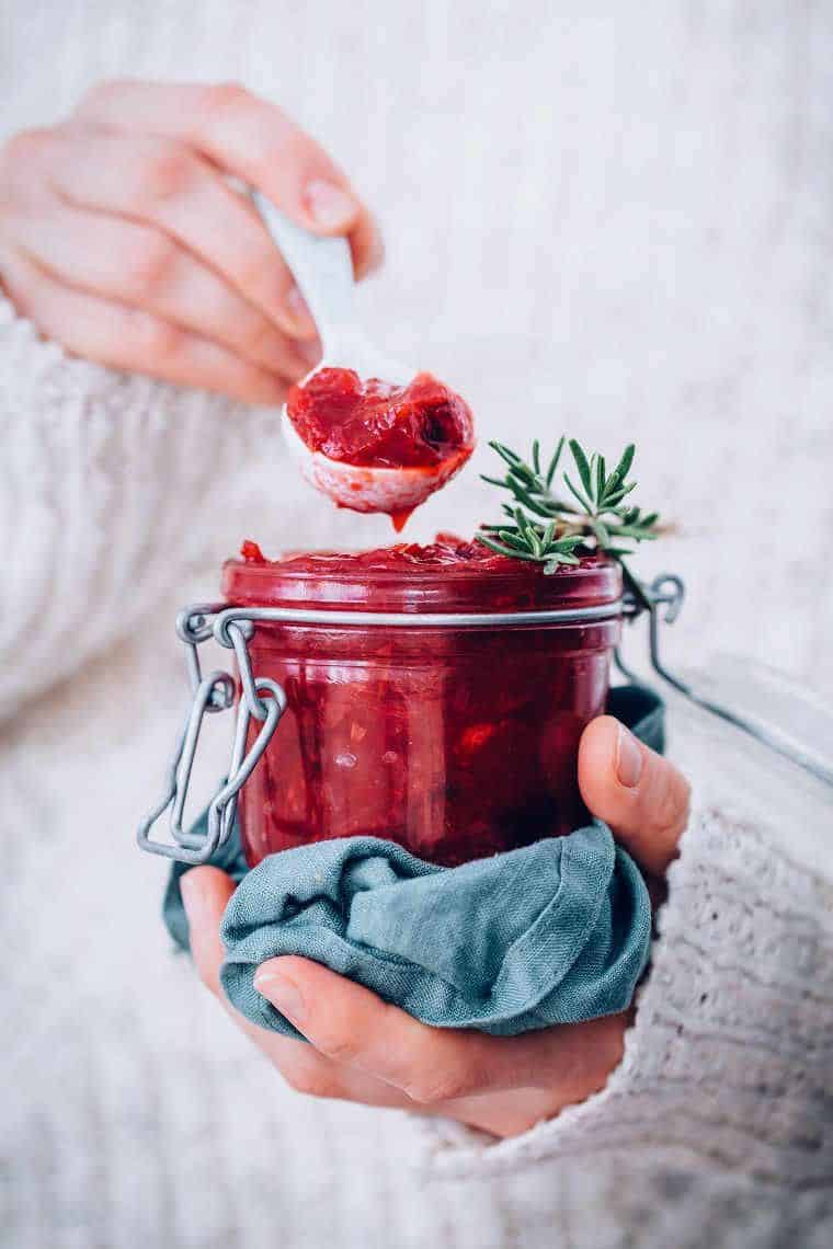 woman in cozy white sweater holding a glass jar with red cranberry chutney and a blue towel as well as a white spoon with some of the chutney