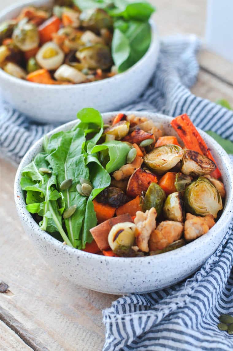 white bowl with roasted carrot, brussels sprouts, tempeh, leafy greens and pumpkin seeds on a white and blue towel