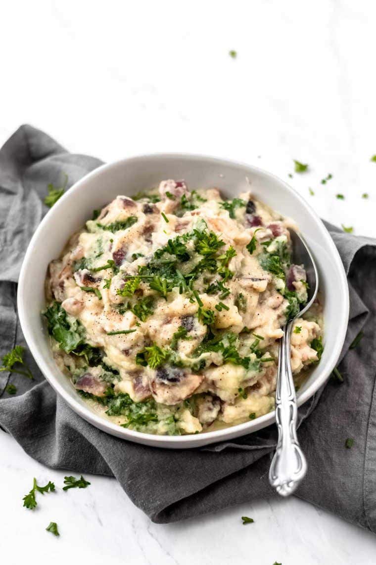 white bowl of creamy vegan mashed potatoes with spinach and mushrooms topped with parsley on a grey towel