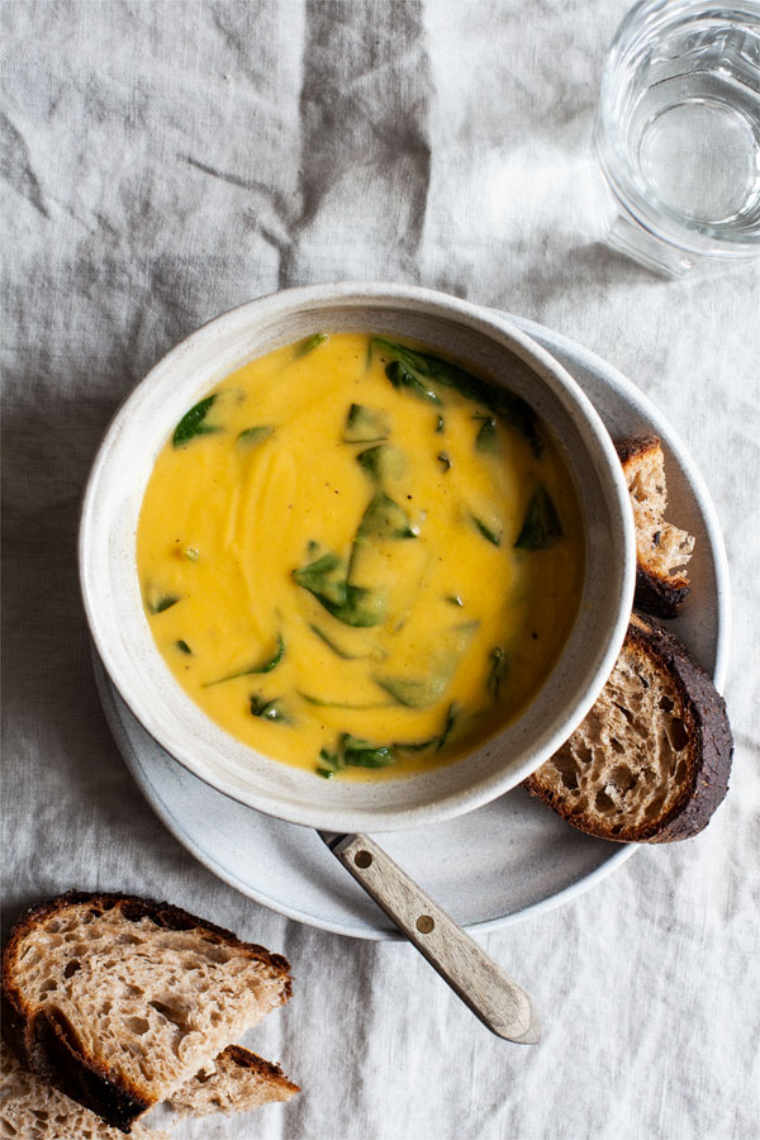 white bowl of roasted garlic chickpea soup with wilted greens next to some sourdough bread