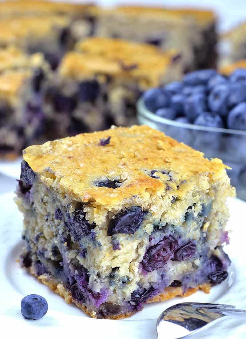 Piece of Blueberry Breakfast Cake on a white plate with a bunch of cakes and blueberries behind.