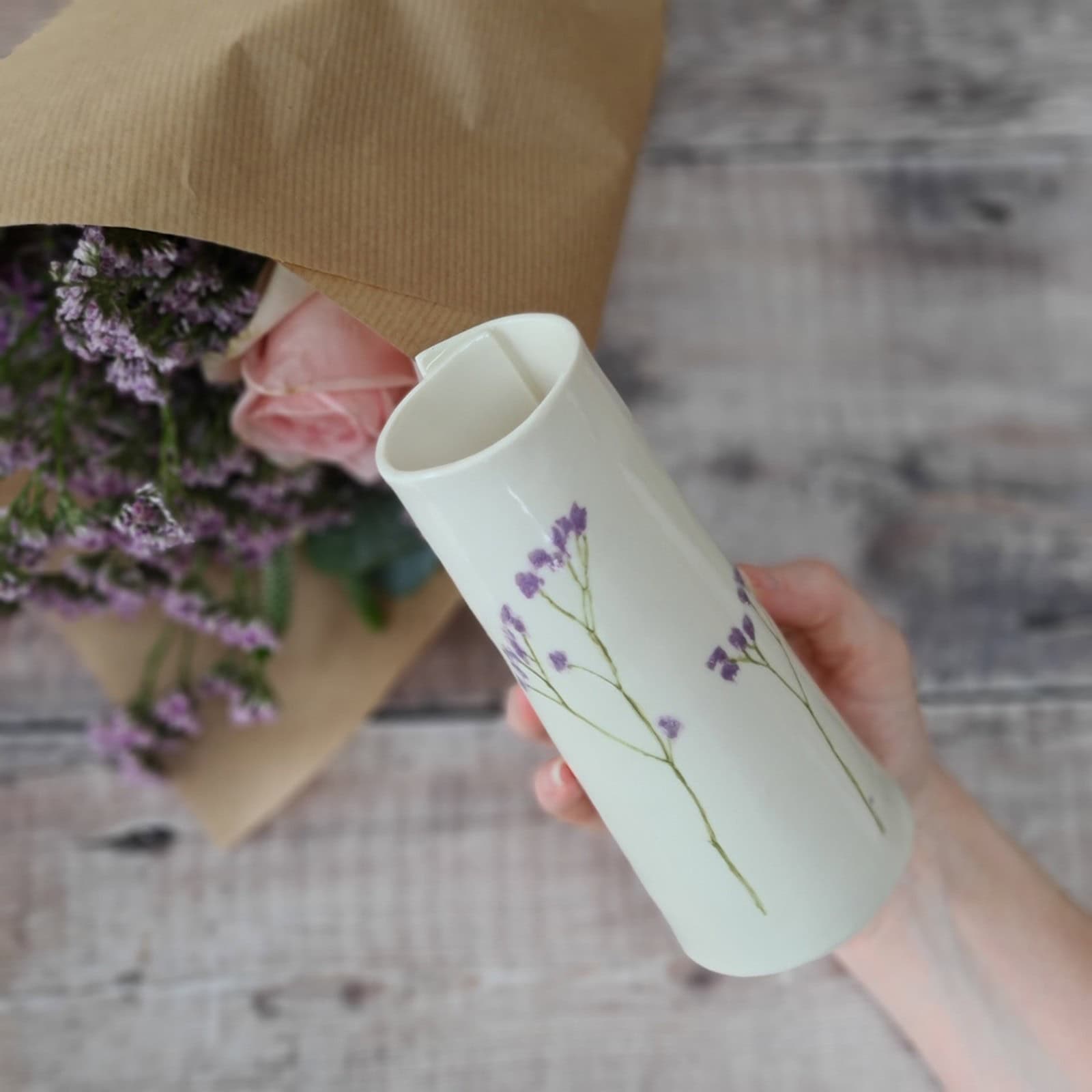 A hand holds a white ceramic vase with purple flowers, next to a wrapped bouquet of pink and purple flowers on a wooden surface.