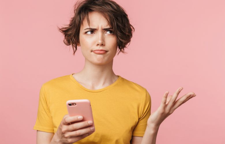 Verwunderte Frau mit Smartphone in der Hand vor rosa Hintergrund – Unsicherheit aufgrund einer schlechten Nutzererfahrung (User Experience) auf einer Praxiswebsite.