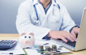 Arzt bei der Buchhaltung am Laptop mit Sparschwein, Münzen und Finanzunterlagen auf dem Schreibtisch.