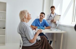 Ein Pfleger und eine Ärztin im aufmerksamen Gespräch mit einer älteren Patientin am Schreibtisch einer hellen Arztpraxis.