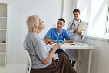 Ein Pfleger und eine Ärztin im aufmerksamen Gespräch mit einer älteren Patientin am Schreibtisch einer hellen Arztpraxis.