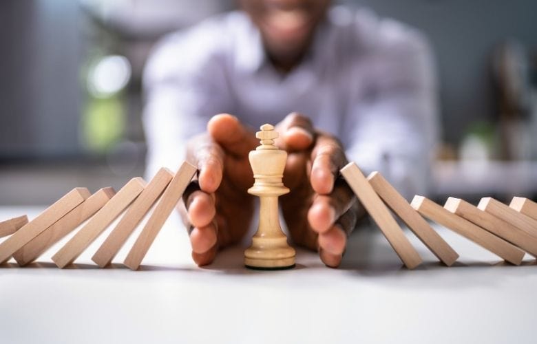 Hände schützen einen weißen Schachkönig vor umfallenden Dominosteinen als Symbol für Abwehr von Gefahren bzw. Risikomanagement in der Arztpraxis