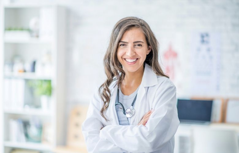 Eine lächelnde Ärztin in weißem Kittel mit Stethoskop und verschränkten Armen in einer hellen Praxisumgebung.