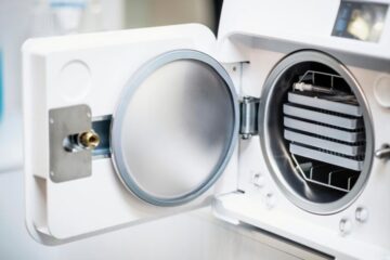 Moderner medizinischer Autoklav mit geöffneter Tür und mehreren metallischen Tabletts zur Instrumentenaufbereitung.