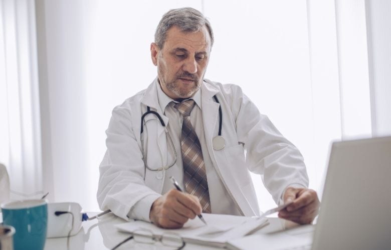 Ein Arzt im weißen Kittel mit Stethoskop sitzt am Schreibtisch und bearbeitet konzentriert Dokumente in seiner Praxis.