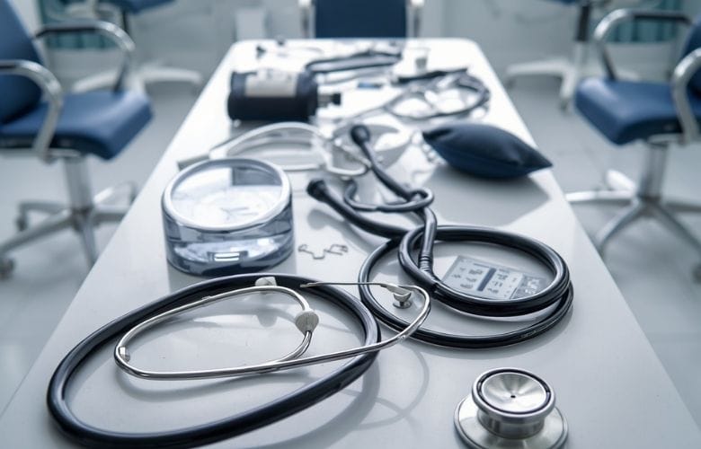 Mehrere Stethoskope und ein Blutdruckmessgerät auf einem hellen Tisch in einer Arztpraxis.