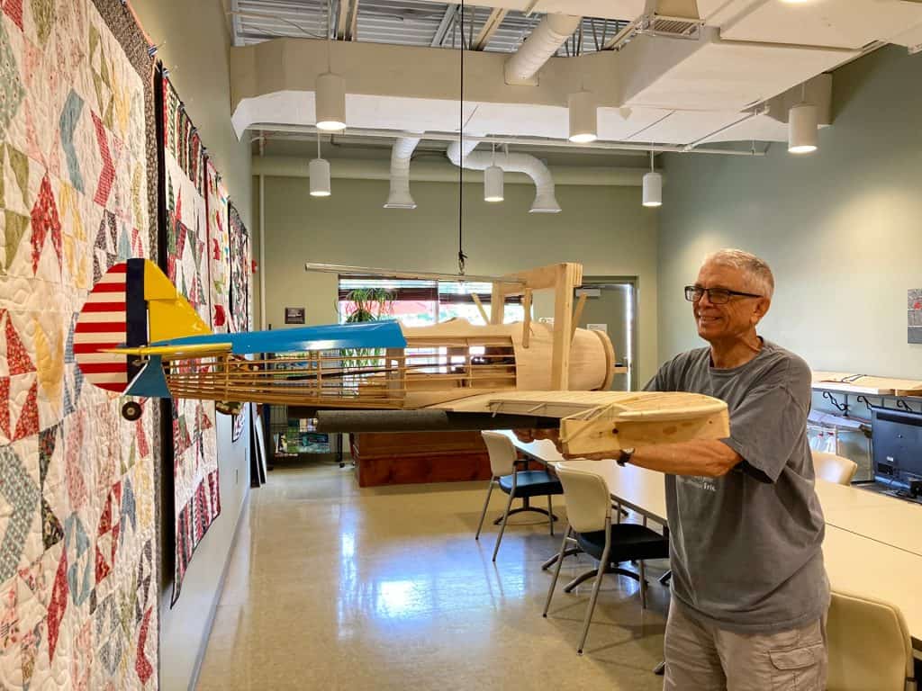 Man holding a large wooden model airplane in a workshop.