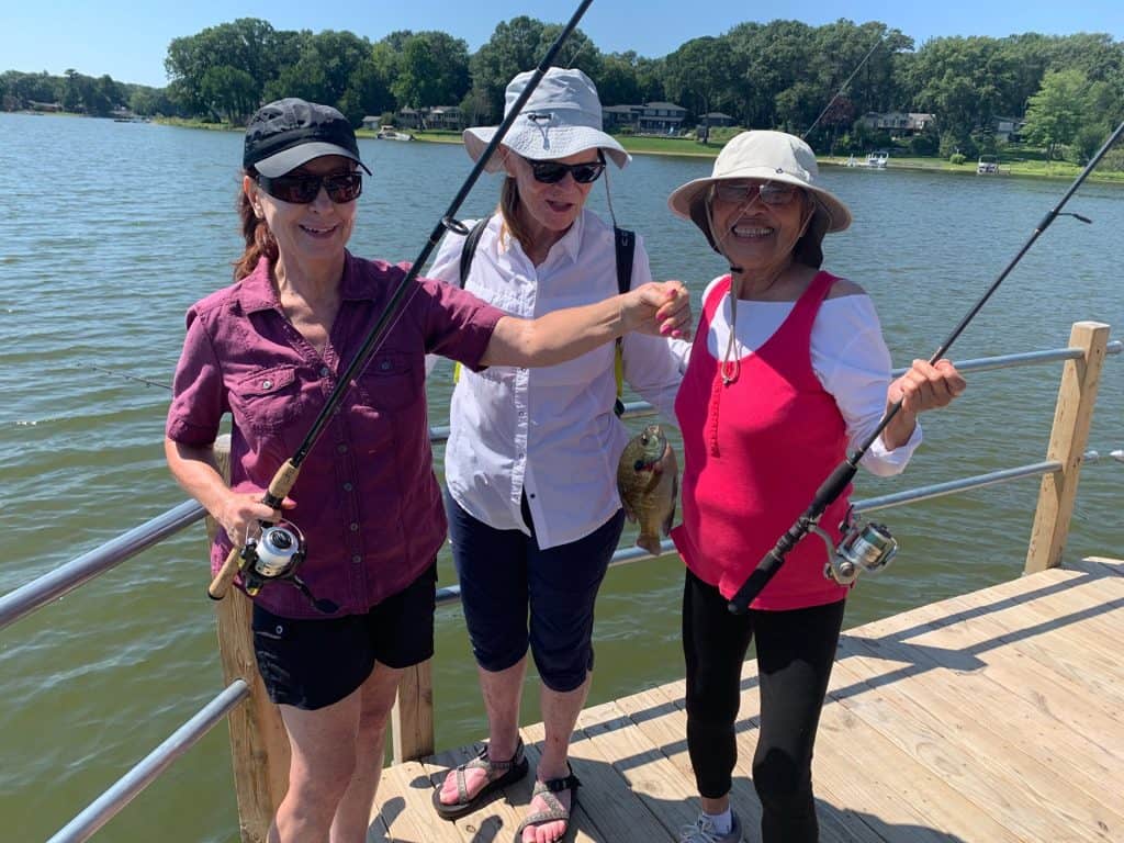 Three women fishing on a wooden dock by a lake with trees in the background.