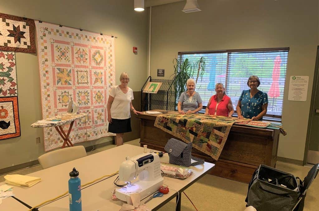 Group of women quilting and displaying finished quilt in a craft room.