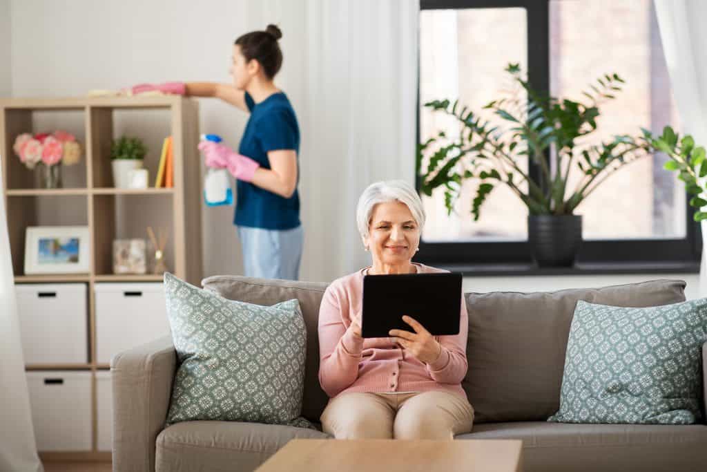 Senior woman using tablet with caregiver cleaning in background.
