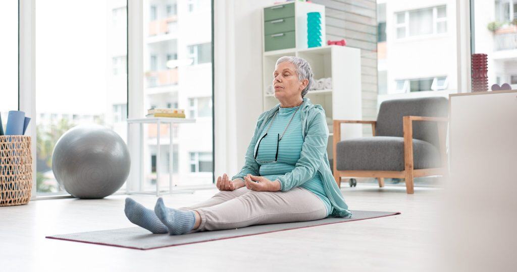 Elderly woman meditating in a bright, modern living room for mindfulness and relaxation.