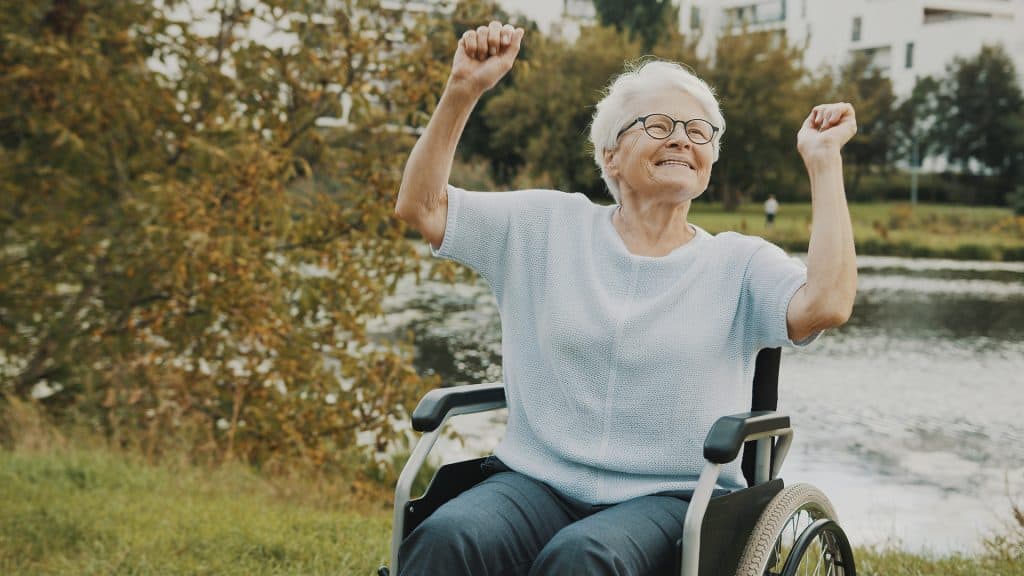 Happy elderly woman in wheelchair celebrating outdoors near water and trees.