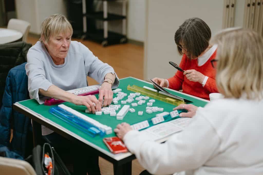 Senior women playing mahjong game at home with tiles and score sheets.