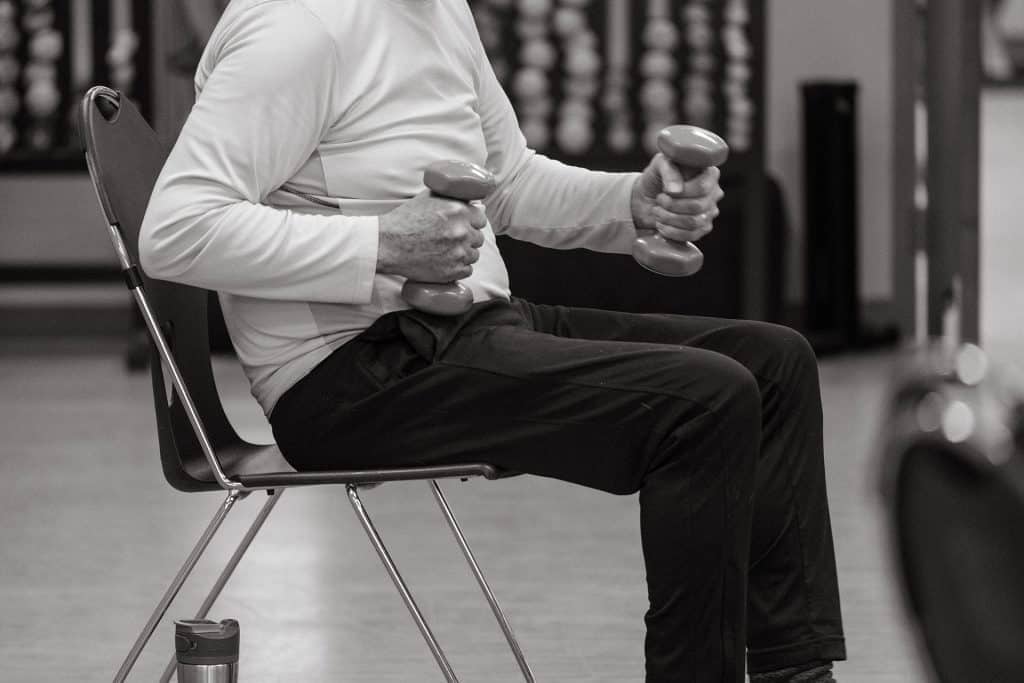Elderly man performing seated strength training with dumbbells in a gym.
