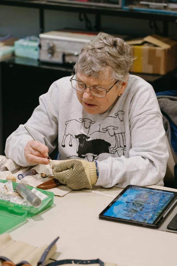 Elderly woman with glasses working on a craft project at a table.