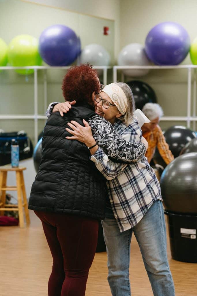 Two senior women hugging in a fitness center with exercise balls in background.