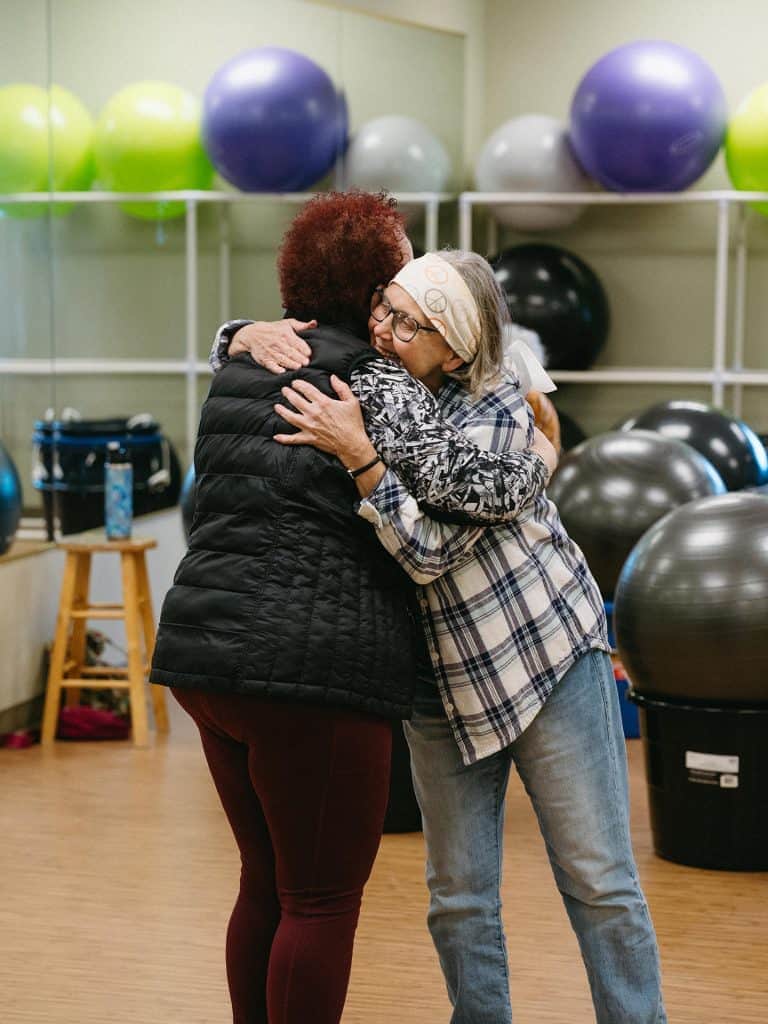 Elderly women hugging in fitness center with exercise balls in background.