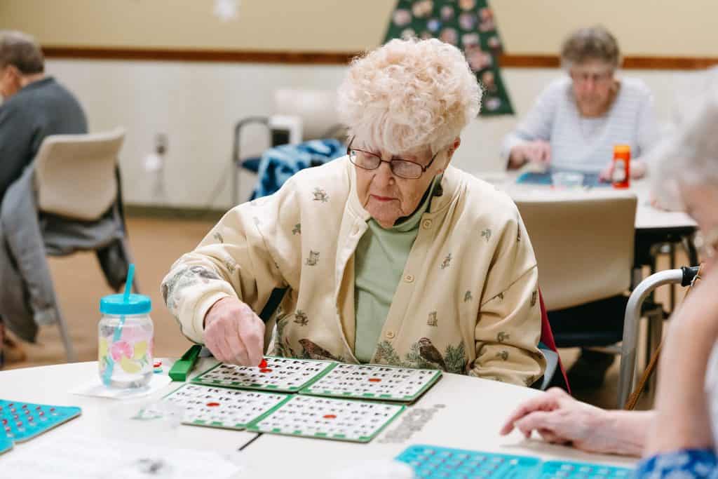 Elderly woman playing bingo in a senior care center with other residents in the background.