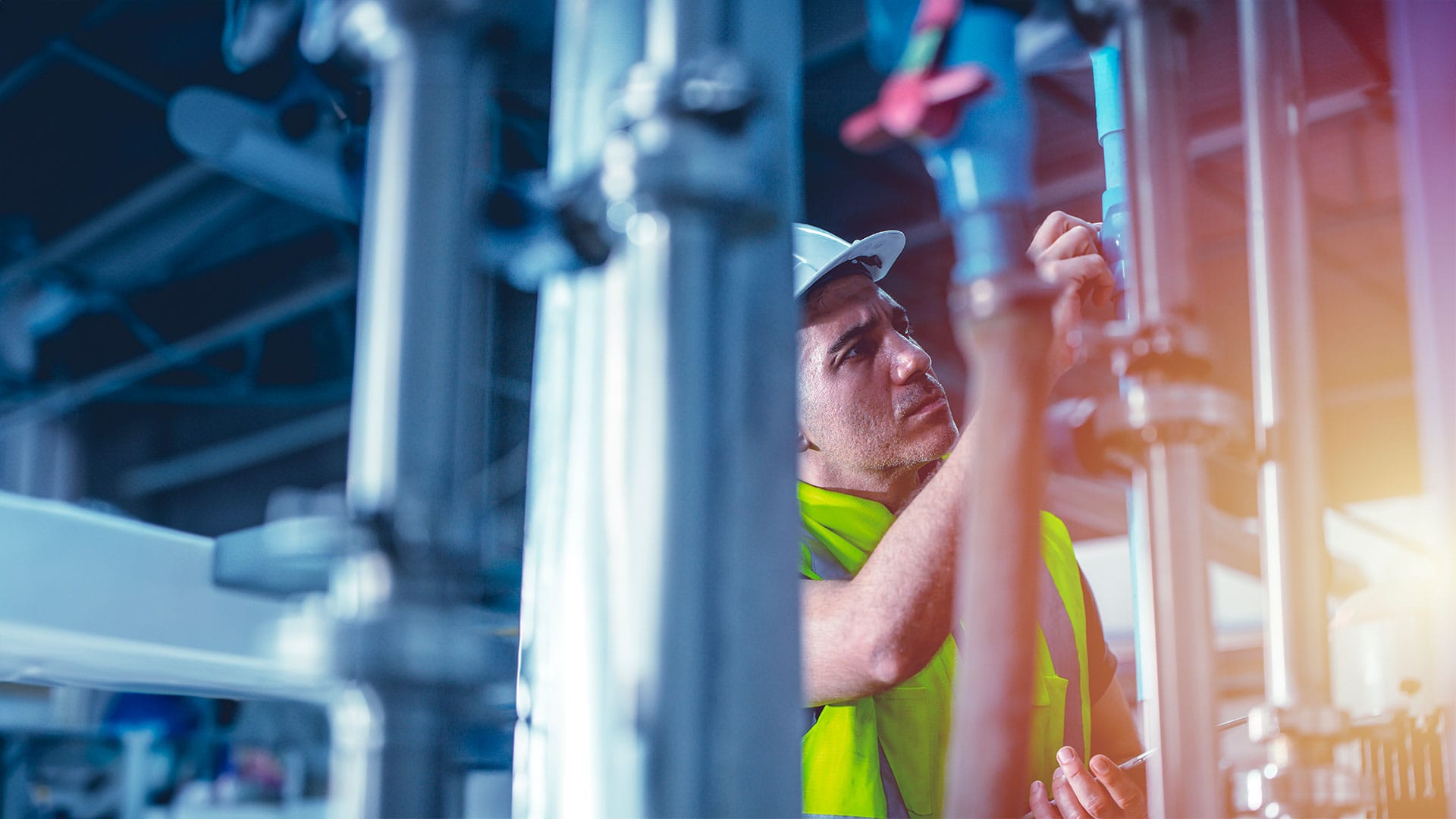 Tecnico in abbigliamento di sicurezza (elmetto bianco e gilet ad alta visibilità) ispeziona un complesso sistema di tubature industriali in un impianto. L'immagine simboleggia innovazione, manutenzione e know-how tecnologico di Nepta e Gruppo Italgas nel settore idrico.