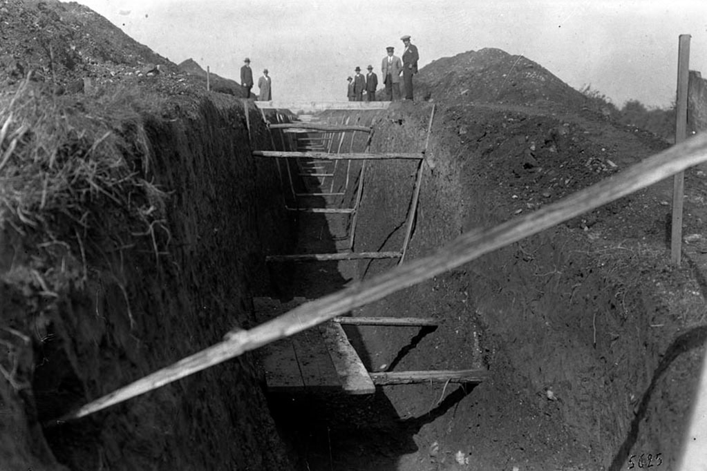 Foto in bianco e nero che mostra una trincea in costruzione, con supporti di legno sopra. Diverse persone sono in piedi sullo sfondo, osservando il cantiere.